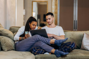 A male and female sitting on the couch in loungewear while on a laptop.