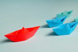A red paper boat in front of two blue paper boats