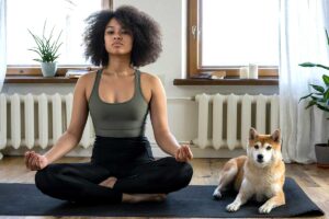 African American woman sitting on the floor doing yoga with her dog next to her.